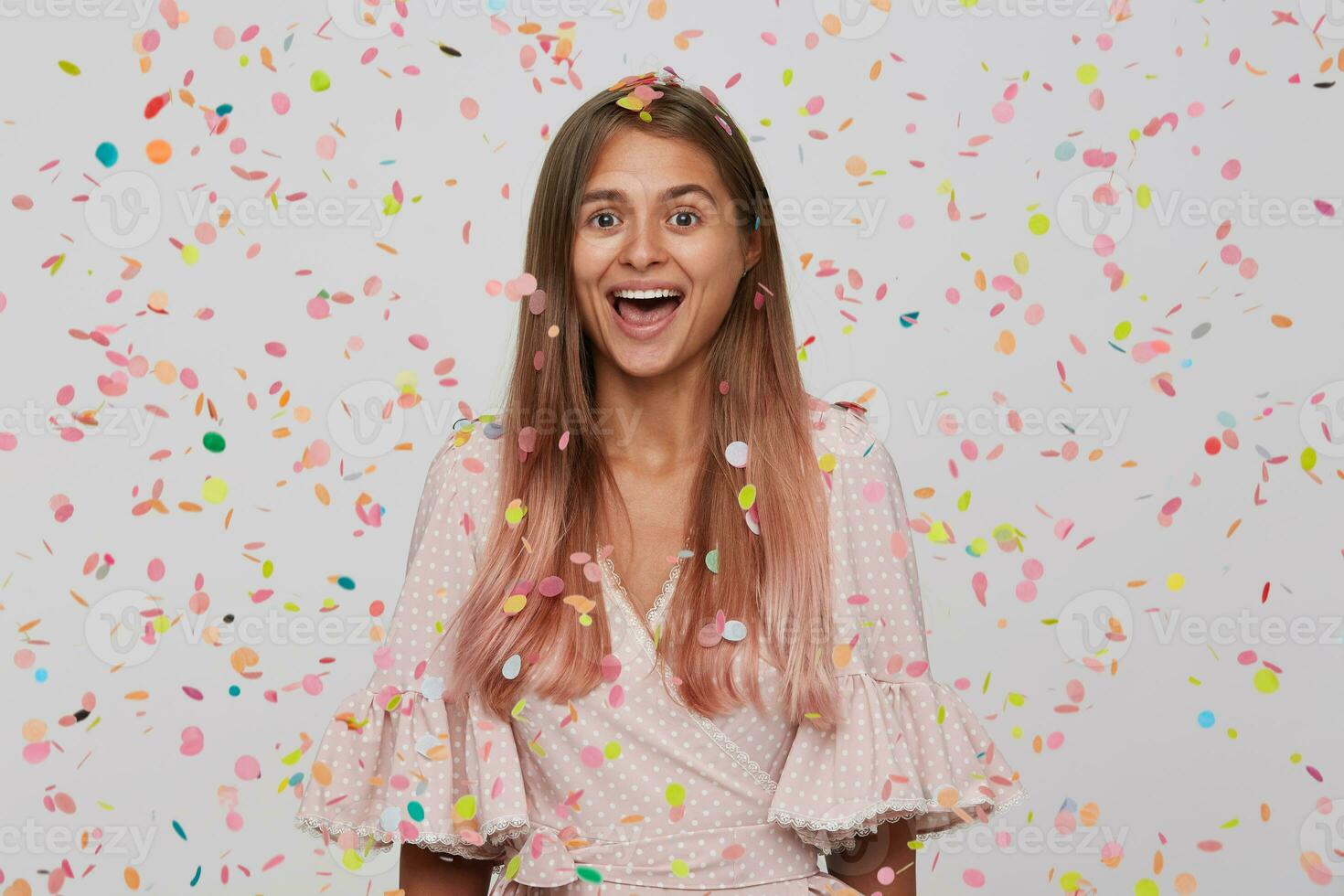 Portrait of cheerful beautiful young woman with long dyed pastel pink hair wears polka dot pink dress feels excited, having party and shouting isolated over white background with confetti photo