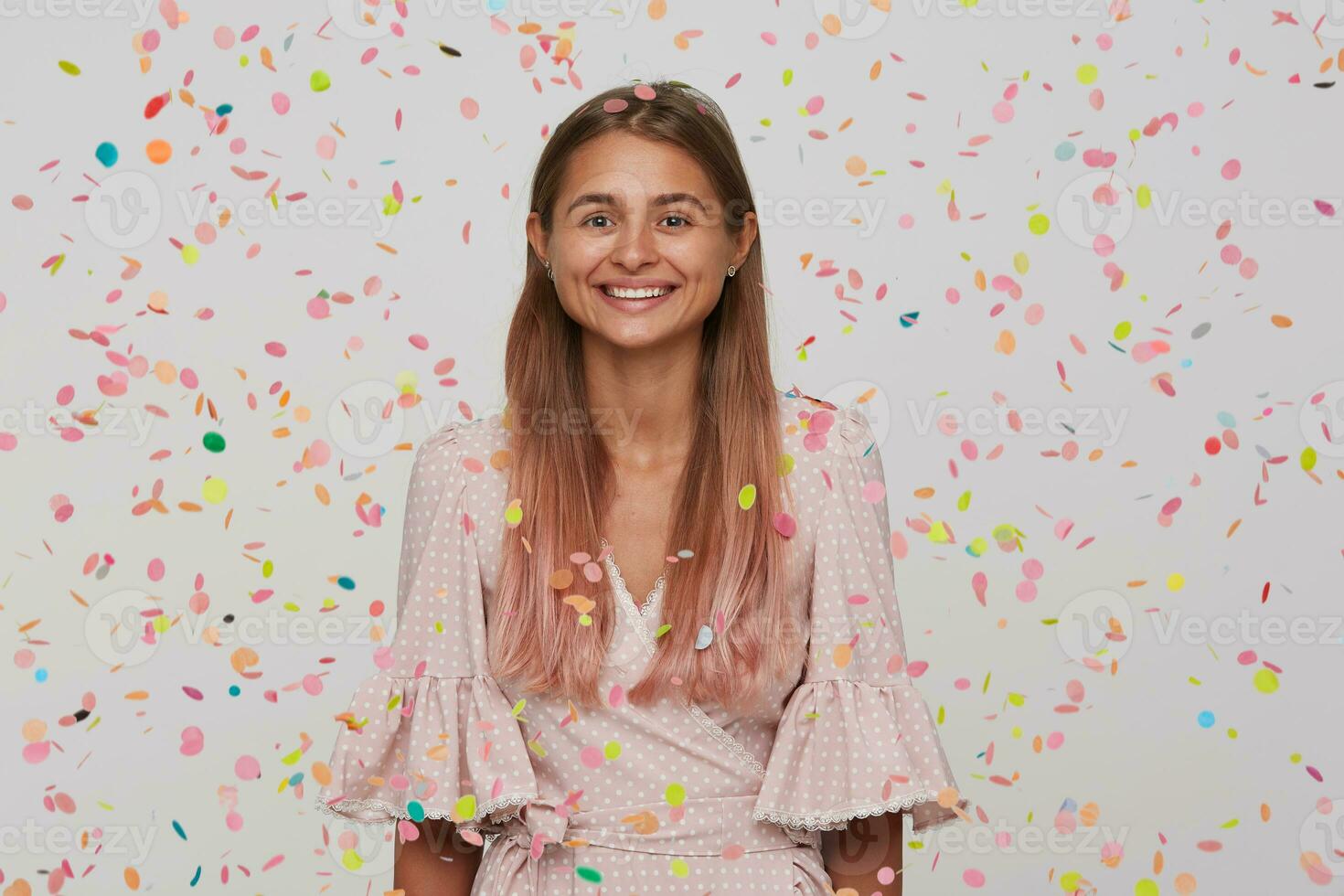 de cerca de alegre atractivo joven mujer con largo teñido pastel rosado pelo usa polca punto rosado vestir teniendo divertido y celebrando cumpleaños aislado terminado blanco antecedentes con papel picado foto