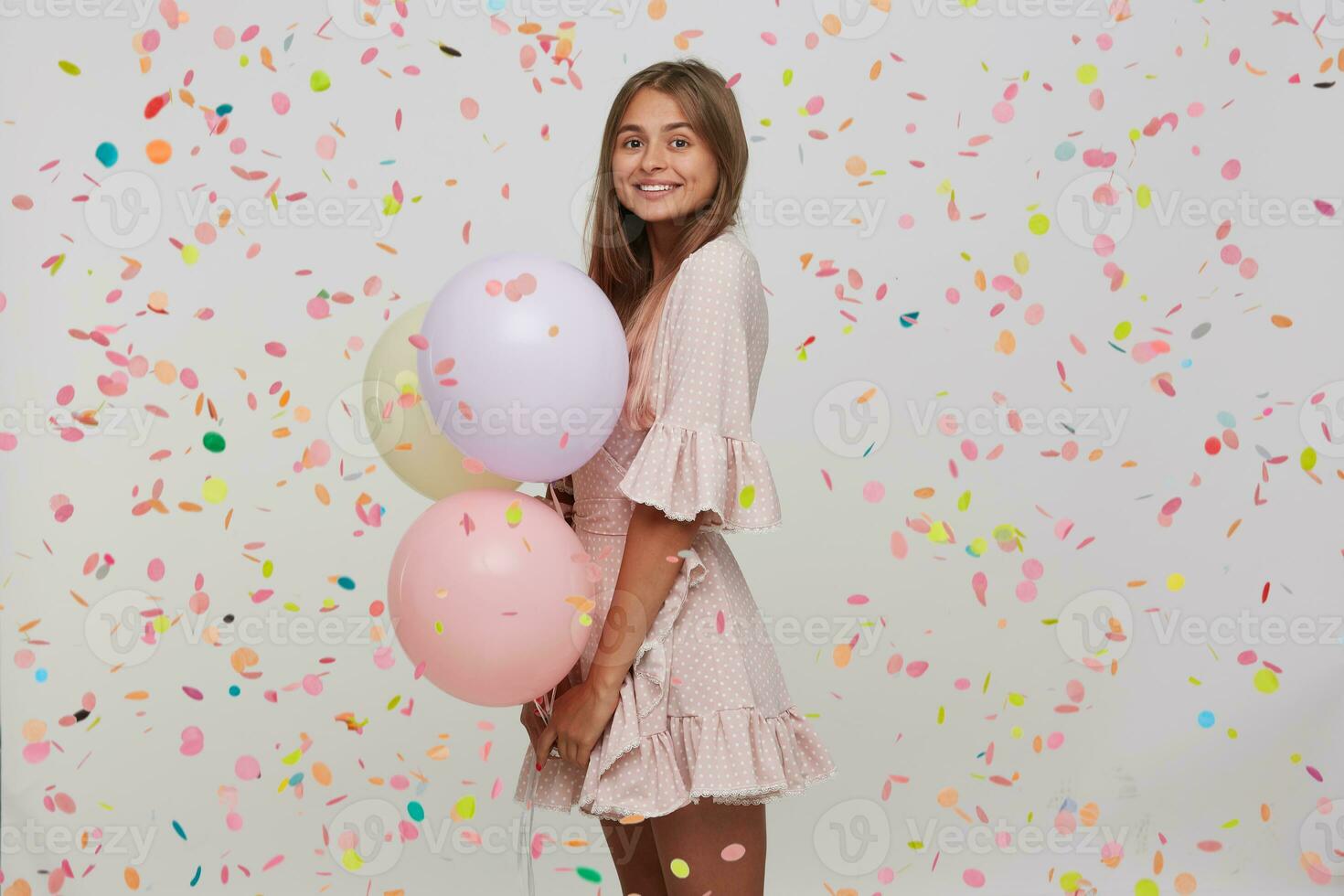 Portrait of happy attractive young woman with long dyed pastel pink hair wears polka dot pink dress holding colorful baloons in hand and having party isolated over white background with confetti photo