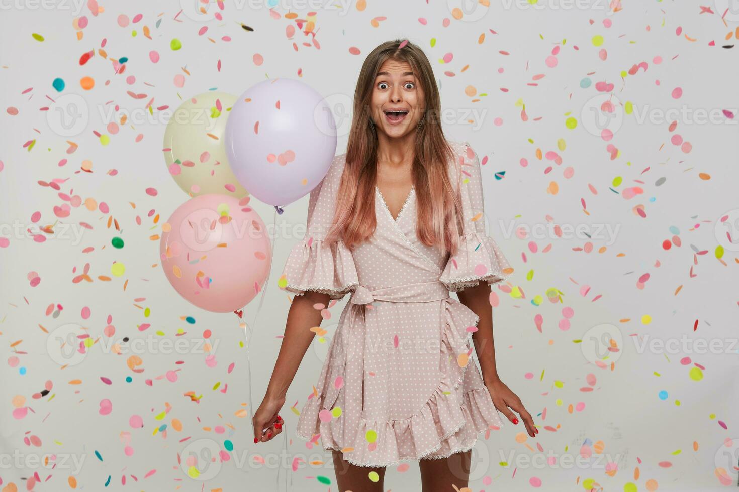 young woman with long dyed pastel pink hair and opened mouth wears polka dot pink dress celebrating birthday, holding colorful baloons in hand isolated over white background with confetti photo