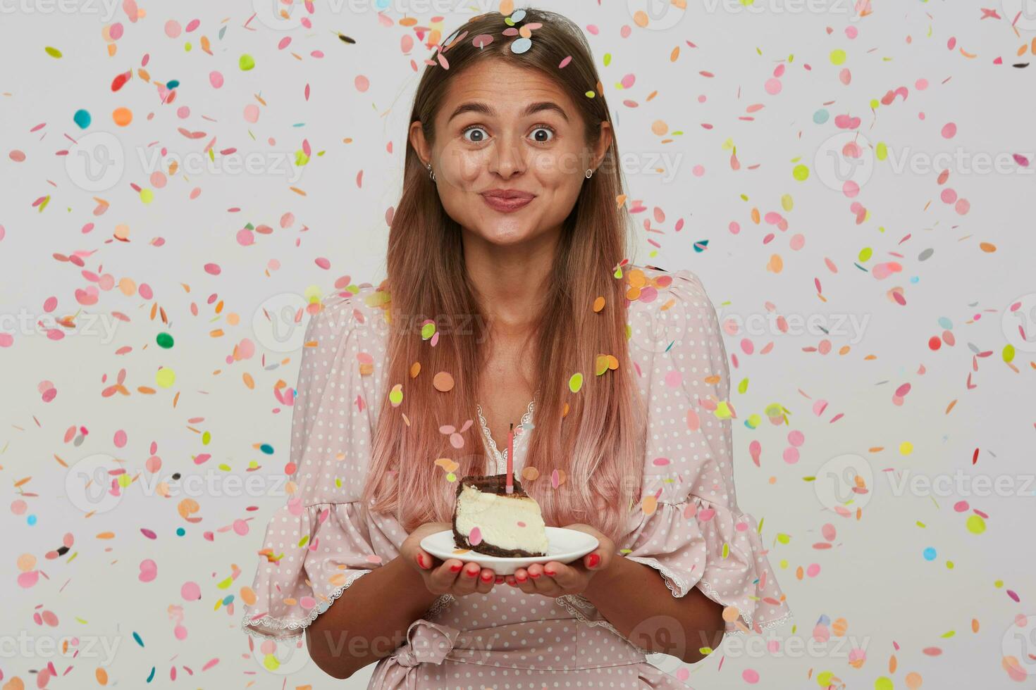 Happy excited young woman with long dyed pastel pink hair wears polka dot pink dress holding plate with piece of cake with candle and having party isolated over white background with confetti photo