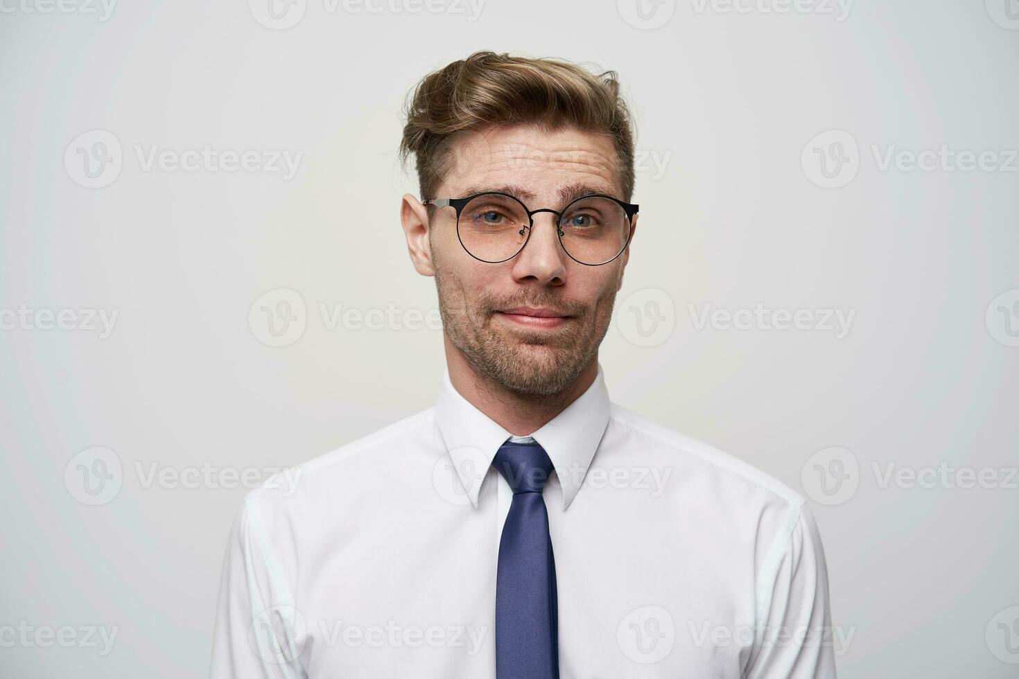 Friendly young man listens attentively, looks calm, with a normal facial expression, works as a consultant or a store manager. Wears white shirt and blue tie, in glasses, over white background photo