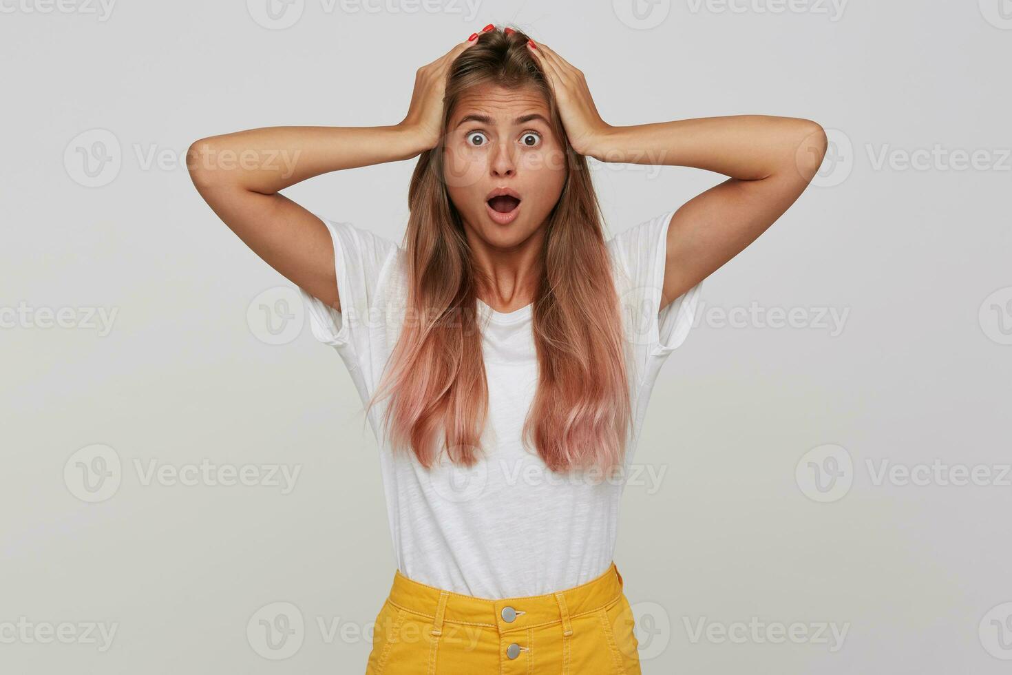 Portrait of amazed stunned young woman with long dyed pastel pink hair and opened mouth keeps hands on head and shouting isolated over white background photo