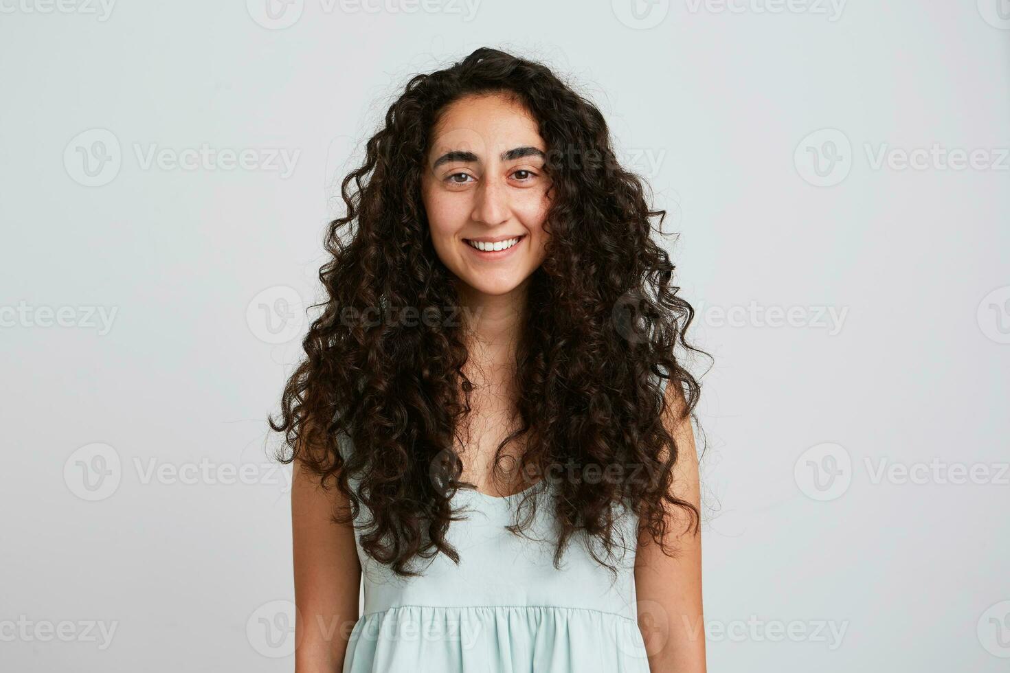 retrato de contento hermosa joven mujer con largo oscuro Rizado pelo sonriente y mira satisfecho aislado terminado blanco antecedentes siente confidente y mirando directamente en cámara foto