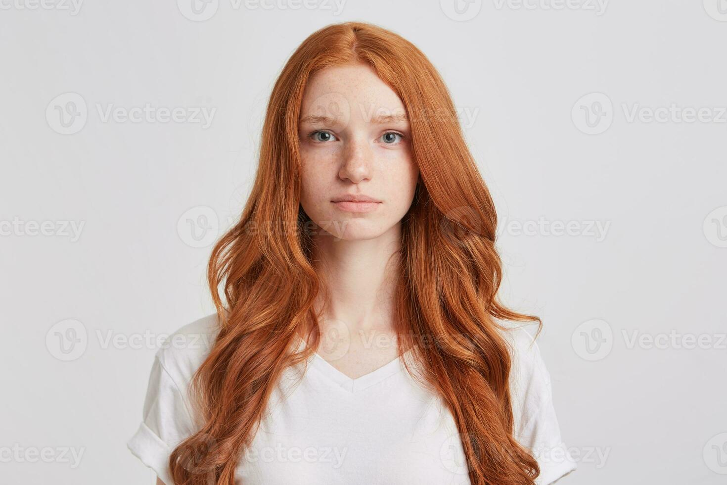 Closeup of serious beautiful redhead young woman with long wavy hair and freckles wears t shirt feels confident and looks directly in camera isolated over white background photo