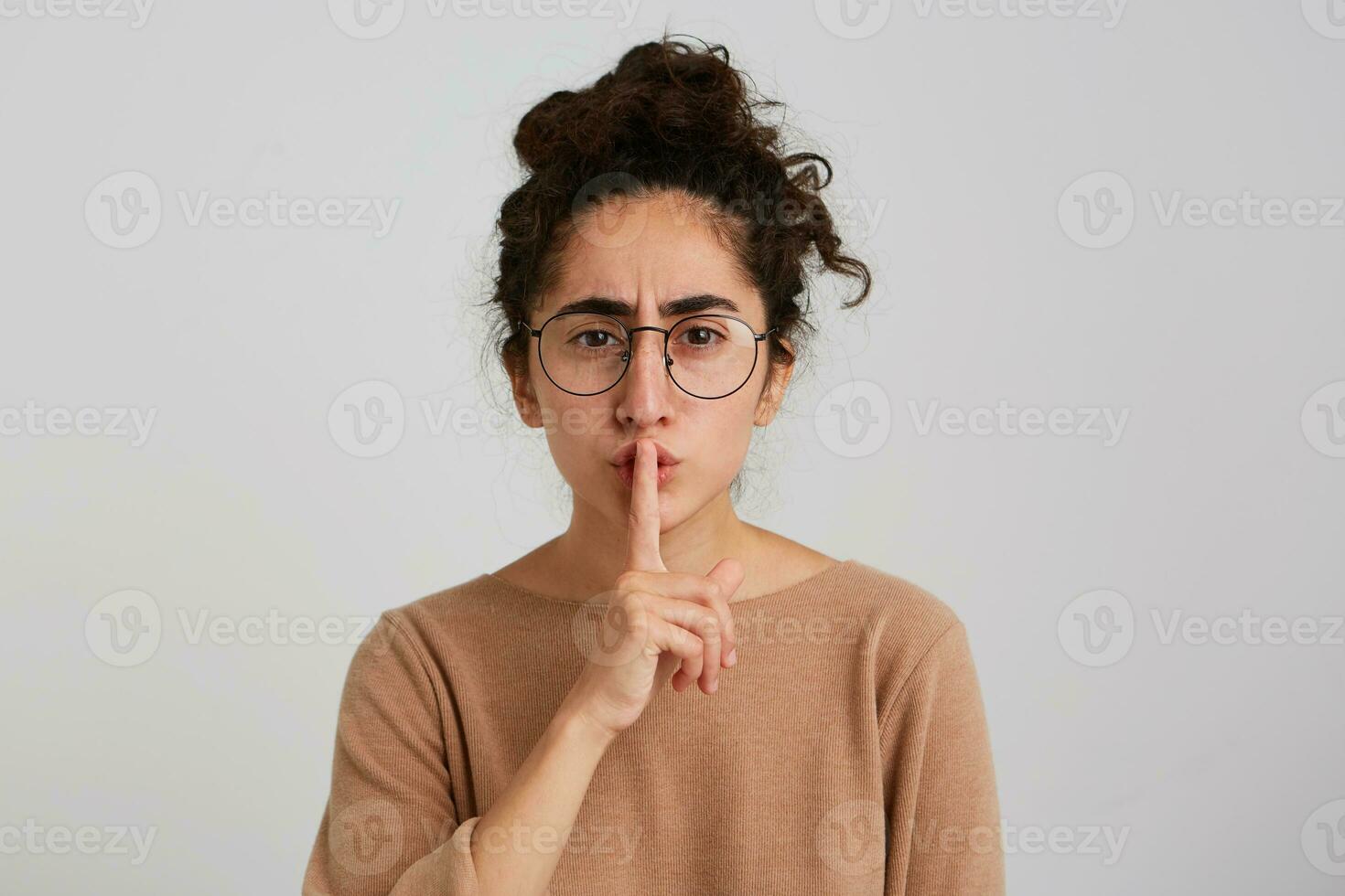 Serious worried young woman with bun of dark curly hair wears beige sweatshirt and glasses looks concerned and shows silence sign isolated over white background photo