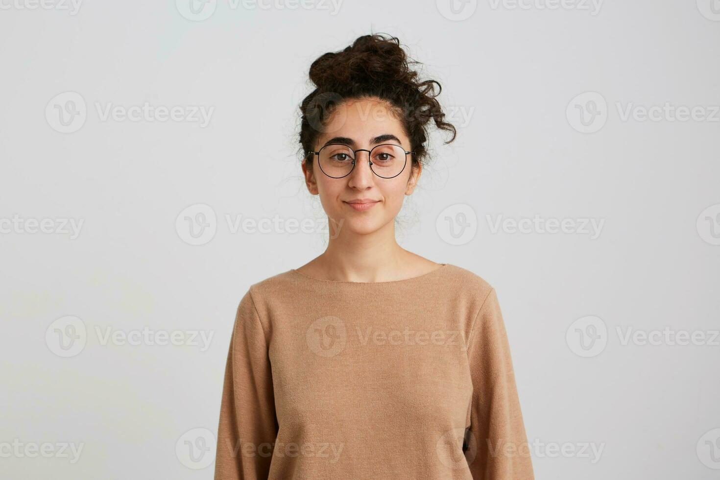 confident pretty young woman student with bun of dark curly hair wears beige pullover and glasses feels peaceful and looks directly in camera isolated over white wall Looking serious and calm photo