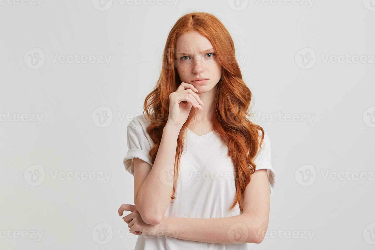 Portrait of pensive worried young woman with long wavy red hair and freckles keeps hands folded and thinking about her problems isolated over white background photo