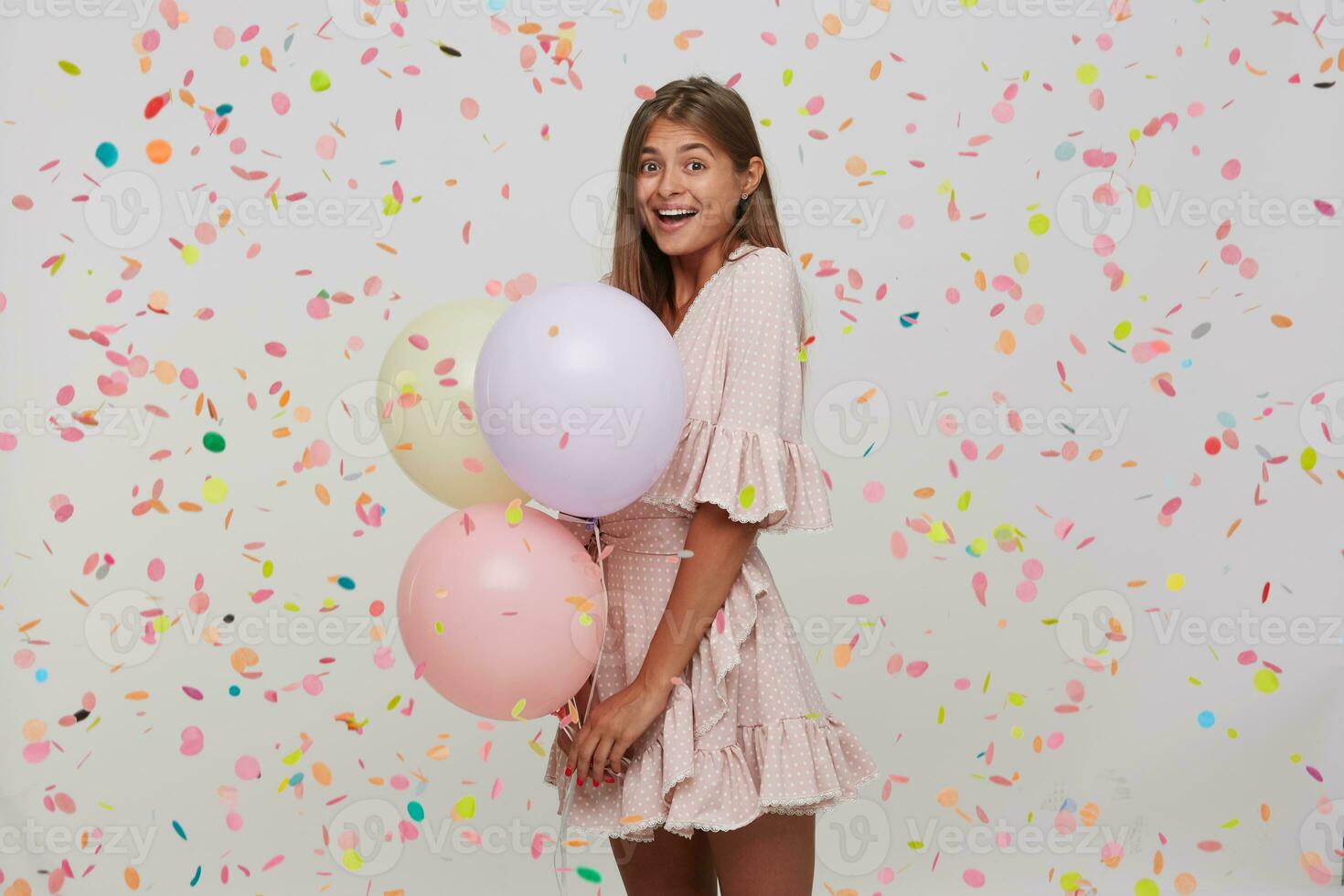 Happy amazed young woman with long hair and opened mouth wears polka dot pink dress celebrating birthday, holding colorful baloons and feels excited isolated over white background with confetti photo