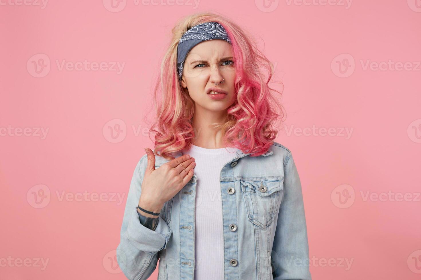 Frowning young cute lady with pink hair, aggressively accepts criticism and looks displeased, points to himself and looks at the camera with disgust, stands over pink background. photo