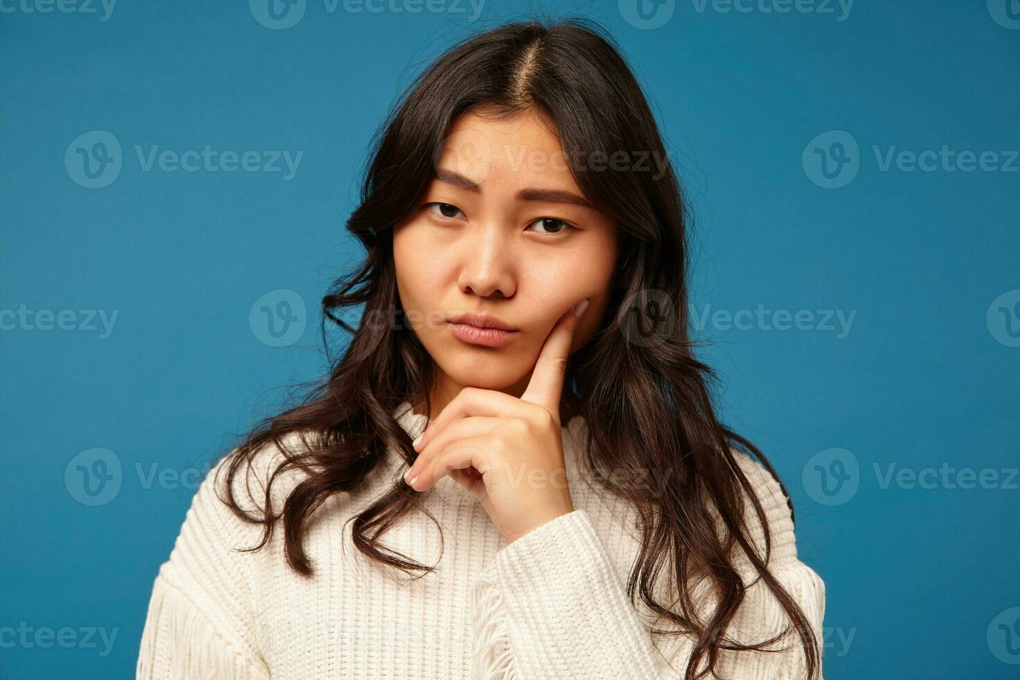Pensive young pretty long haired asian female with natural makeup keeping index finger on her cheek while looking thoughtfully at camera, posing over blue background photo
