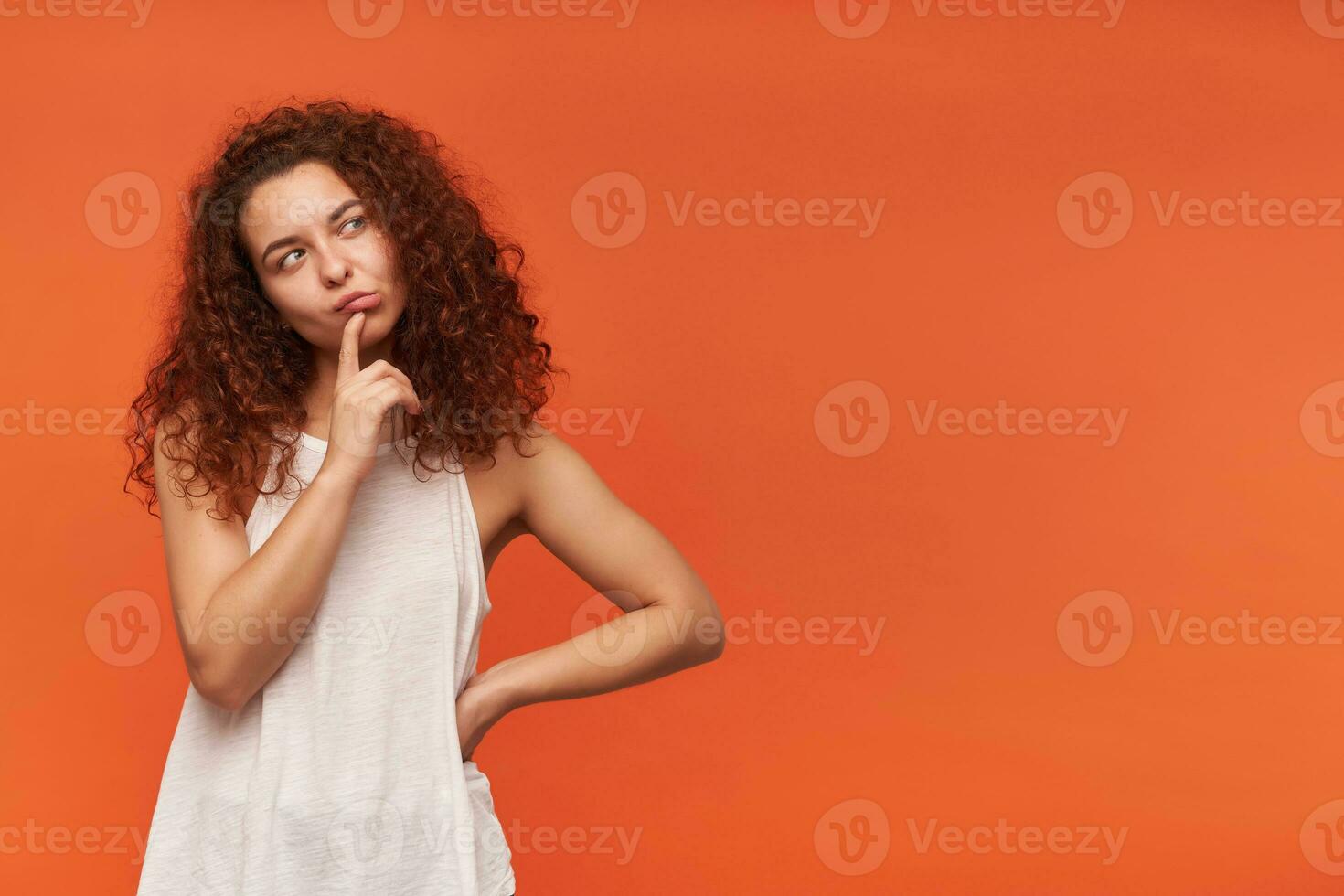 Nice looking woman, beautiful girl with curly ginger hair. Wearing white off-shoulder blouse. Touching her lip and thinking. Watching to the right at copy space, isolated over orange background photo