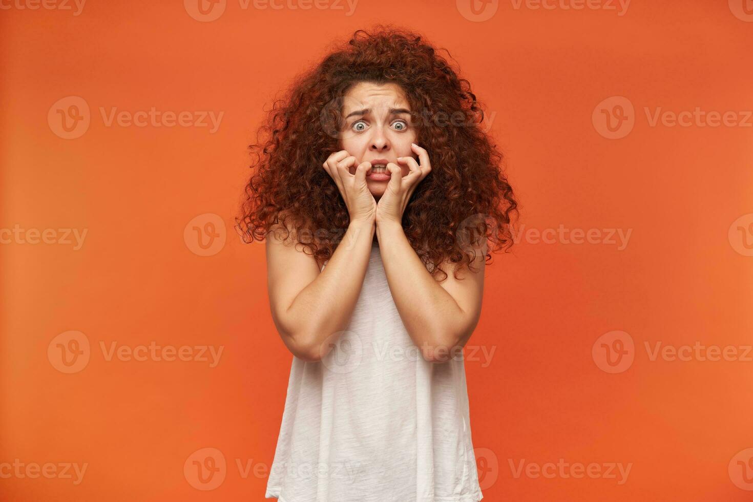 Afraid looking woman, girl with curly ginger hair. Wearing white off-shoulder blouse. Touching her face. Has fear on her face. Watching at the camera horrified, isolated over orange background photo