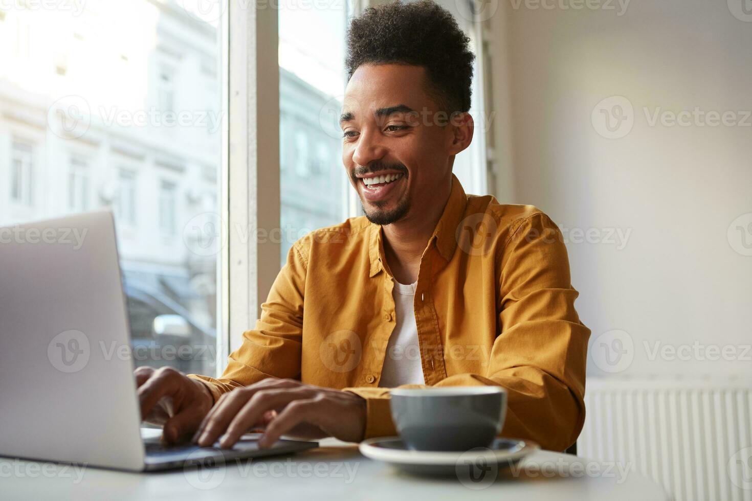 Young African American man sitting at a table in a cafe and working on a laptop, wears yellow shirt, drinks aromatic coffee, creates new content for his blog, enjoys the work of a freelancer. photo