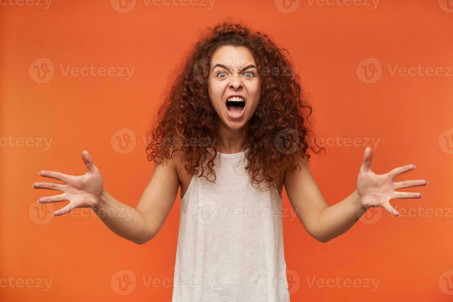 Portrait of evil, adult redhead girl with curly hair. Wearing white off-shoulder blouse. Trying to scare you. Screams and spreading her arms. Watching at the camera, isolated over orange background photo