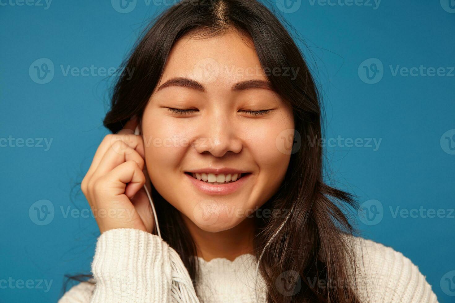 Closeup of young good looking dark haired lady with romantic hairstyle keeping eyes closed while enjoying her favourite music, posing over blue background photo