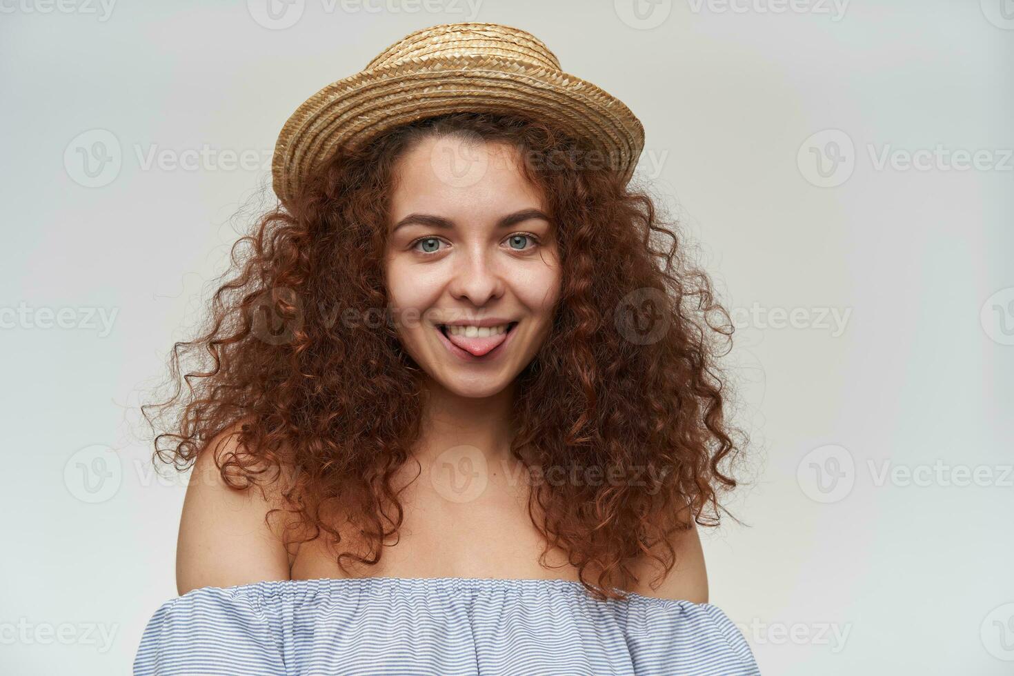 Portrait of attractive, adult redhead girl with curly hair. Wearing striped off-shoulders blouse and hat. Showing tongue. Watching at the camera, closeup, isolated over white background photo