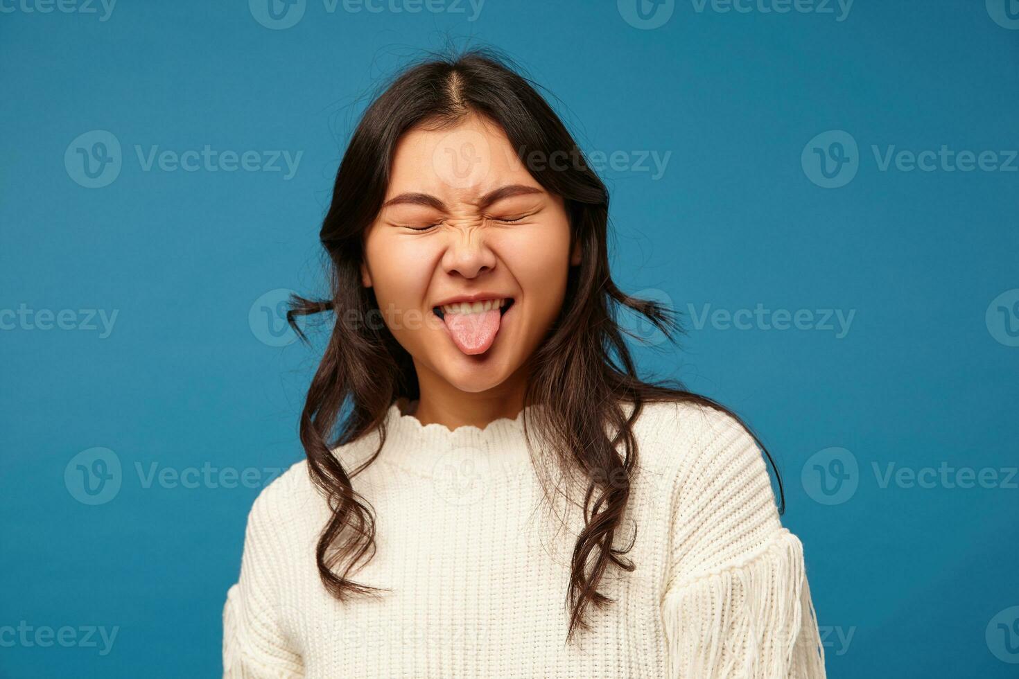 Good looking young pretty long haired woman with natural makeup keeping her eyes closed while making funny faces, showing her tongue while standing over blue background photo