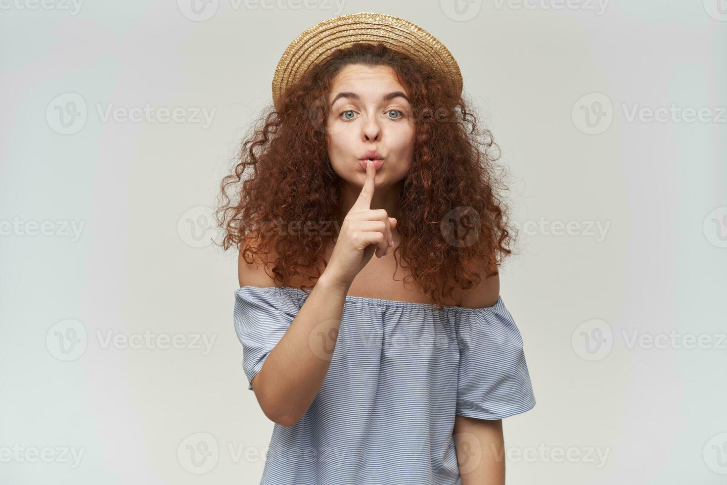 Nice looking woman, beautiful girl with curly ginger hair. Wearing striped off-shoulders blouse and hat. Showing silence sign. Watching at the camera, isolated over white background photo