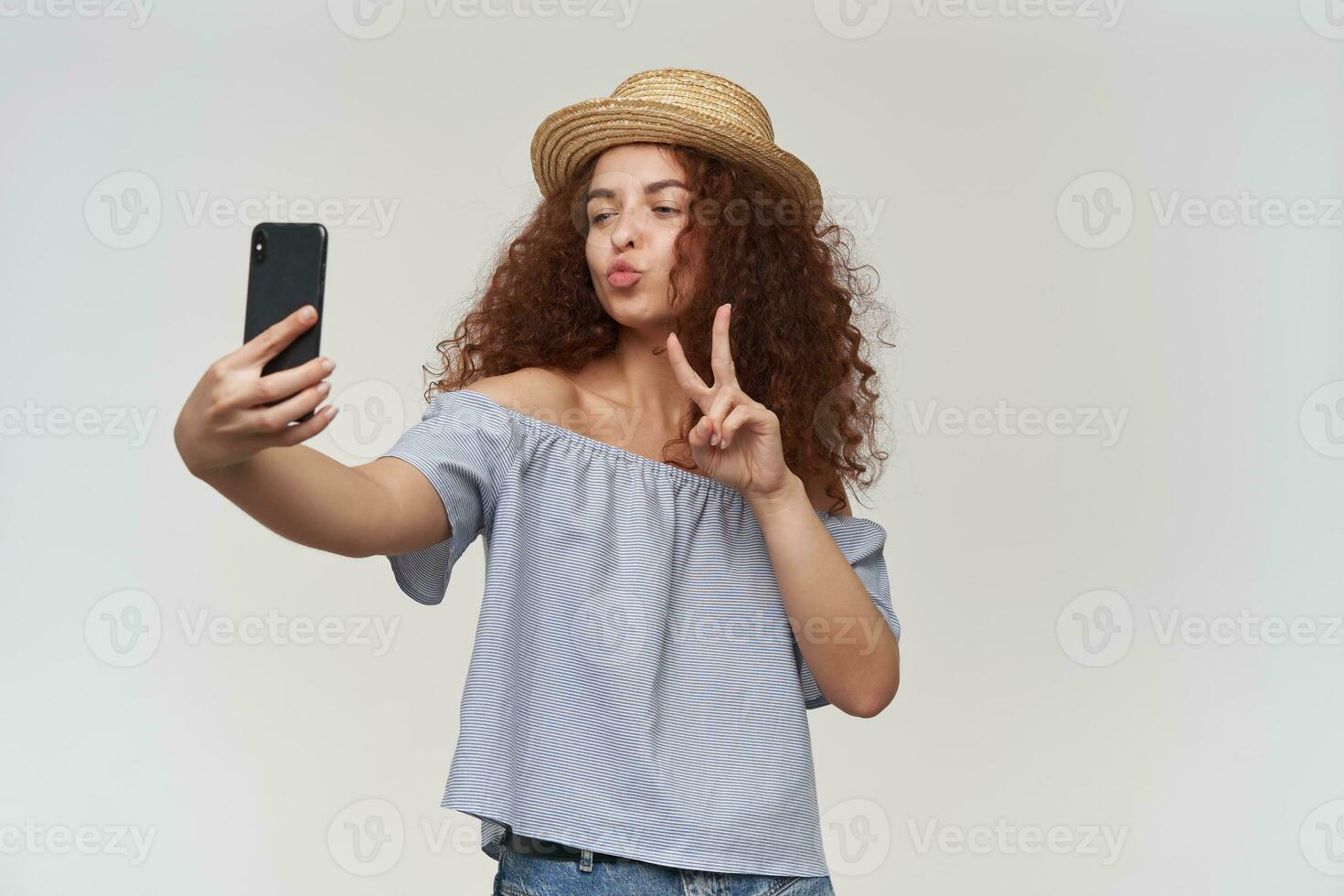 Adolescente chica, contento mujer con Rizado jengibre cabello. vistiendo a rayas fuera de los hombros blusa y sombrero. tomando un selfie en un teléfono inteligente, demostración paz firmar y beso. estar aislado terminado blanco antecedentes foto