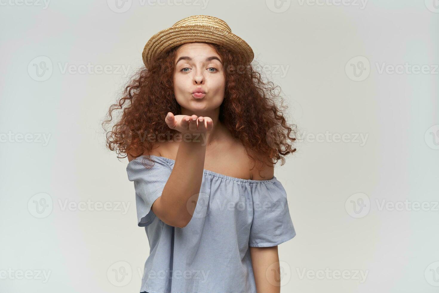 Portrait of attractive, adult redhead girl with curly hair. Wearing striped off-shoulders blouse and hat. Sending air kiss. Watching lovely at the camera, isolated over white background photo