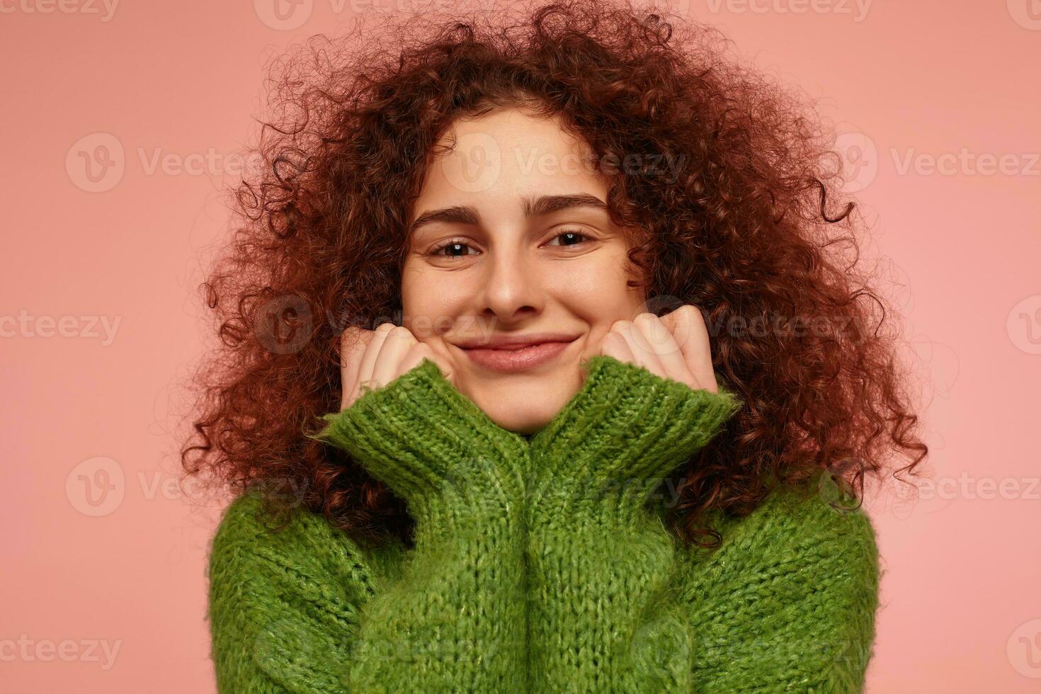 joven dama, bonito mujer con jengibre Rizado cabello. vistiendo verde suéter tipo con cuello de tortuga suéter, siente calentar y conmovedor su las mejillas, sonriente. acecho a el cámara aislado, de cerca terminado pastel rosado antecedentes foto