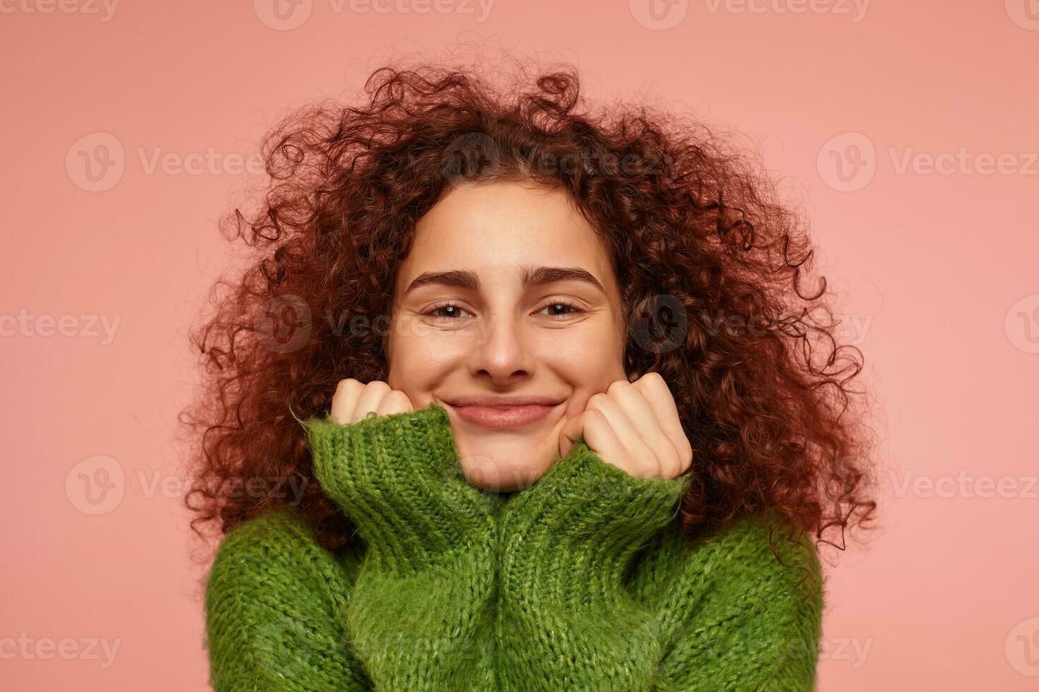 Teenage girl, happy looking redhead woman with curly hair. Wearing green turtleneck sweater and touching her cheeks, smiling. Watching at the camera isolated, closeup over pastel pink background photo