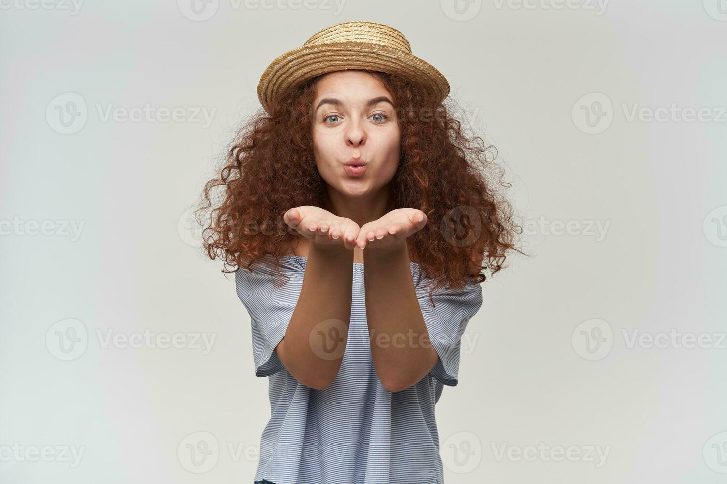 Nice looking woman, beautiful girl with curly ginger hair. Wearing striped off-shoulders blouse and hat. Sending air kiss to you. Watching at the camera, isolated over white background photo