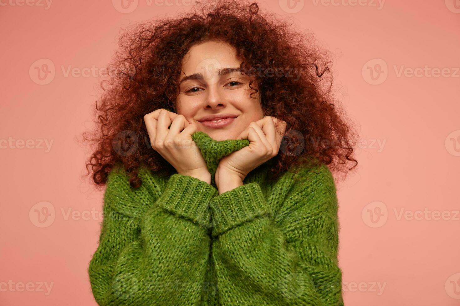 joven dama, bonito mujer con jengibre Rizado cabello. vistiendo verde suéter tipo con cuello de tortuga suéter y Halar un suéter terminado su rostro, sonriente. acecho a el cámara aislado, de cerca terminado pastel rosado antecedentes foto