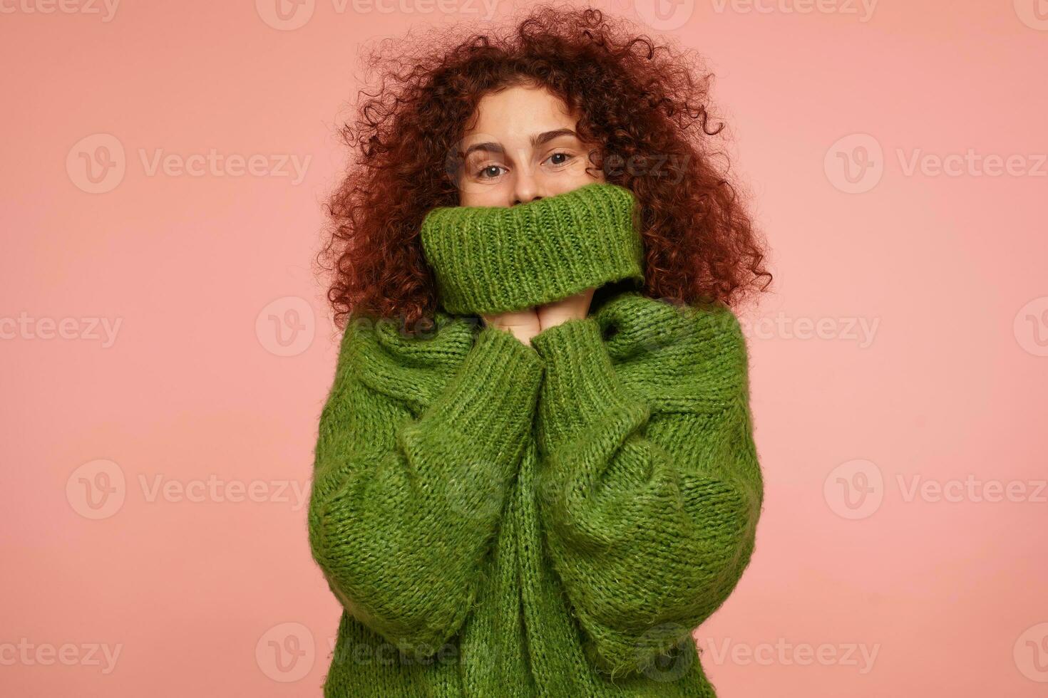 Portrait of attractive, adult girl with ginger curly hair. Wearing green turtleneck sweater and pull a sweater over her face. Watching at the camera isolated over pastel pink background photo