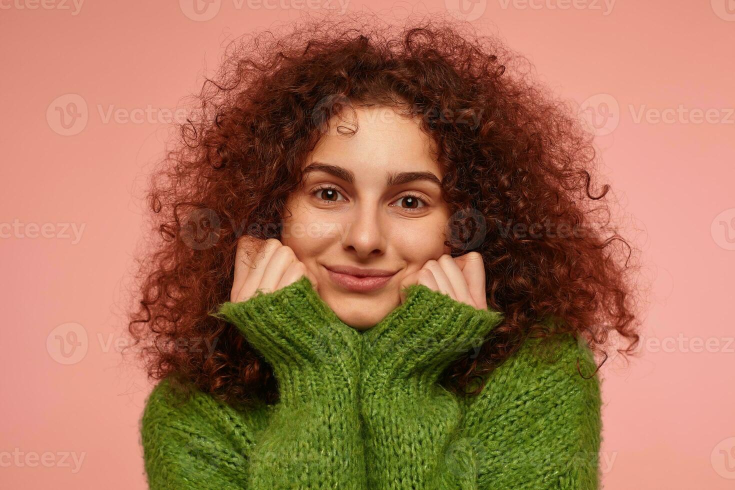 Teenage girl, happy looking redhead woman with curly hair. Wearing green turtleneck sweater and feels warm, touching her cheeks. Watching at the camera isolated, closeup over pastel pink background photo