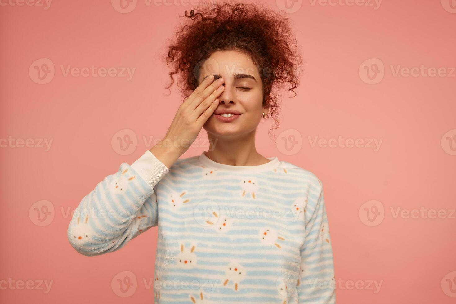 Teenage girl, sleepy looking redhead woman with curly hair. Wearing striped sweater with bunnies and rubs an eye. Morning mood. Stand isolated over pastel pink background photo