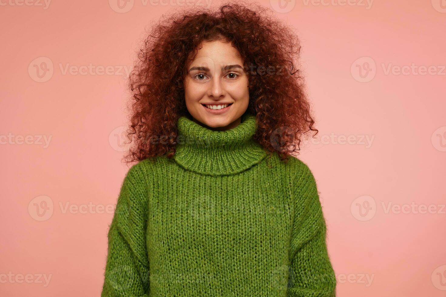 joven dama, bonito mujer con jengibre Rizado cabello. vistiendo verde suéter tipo con cuello de tortuga suéter y tener un genial brillante sonrisa, seguro. acecho a el cámara aislado terminado pastel rosado antecedentes foto