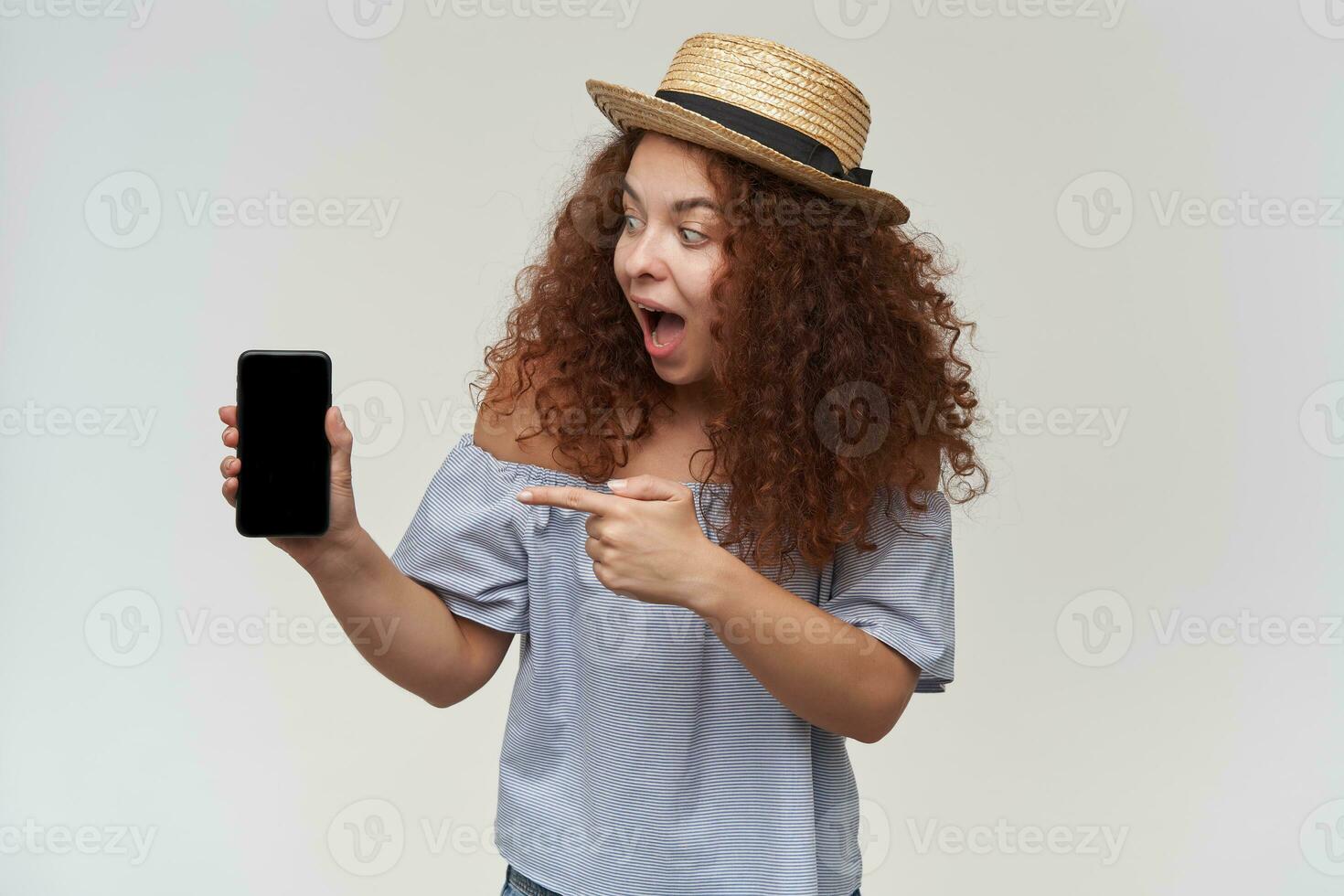 Teenage girl, surprised looking woman with curly ginger hair. Wearing striped off-shoulders blouse and hat. Holding and pointing on her phone, copy space. Stand isolated over white background photo
