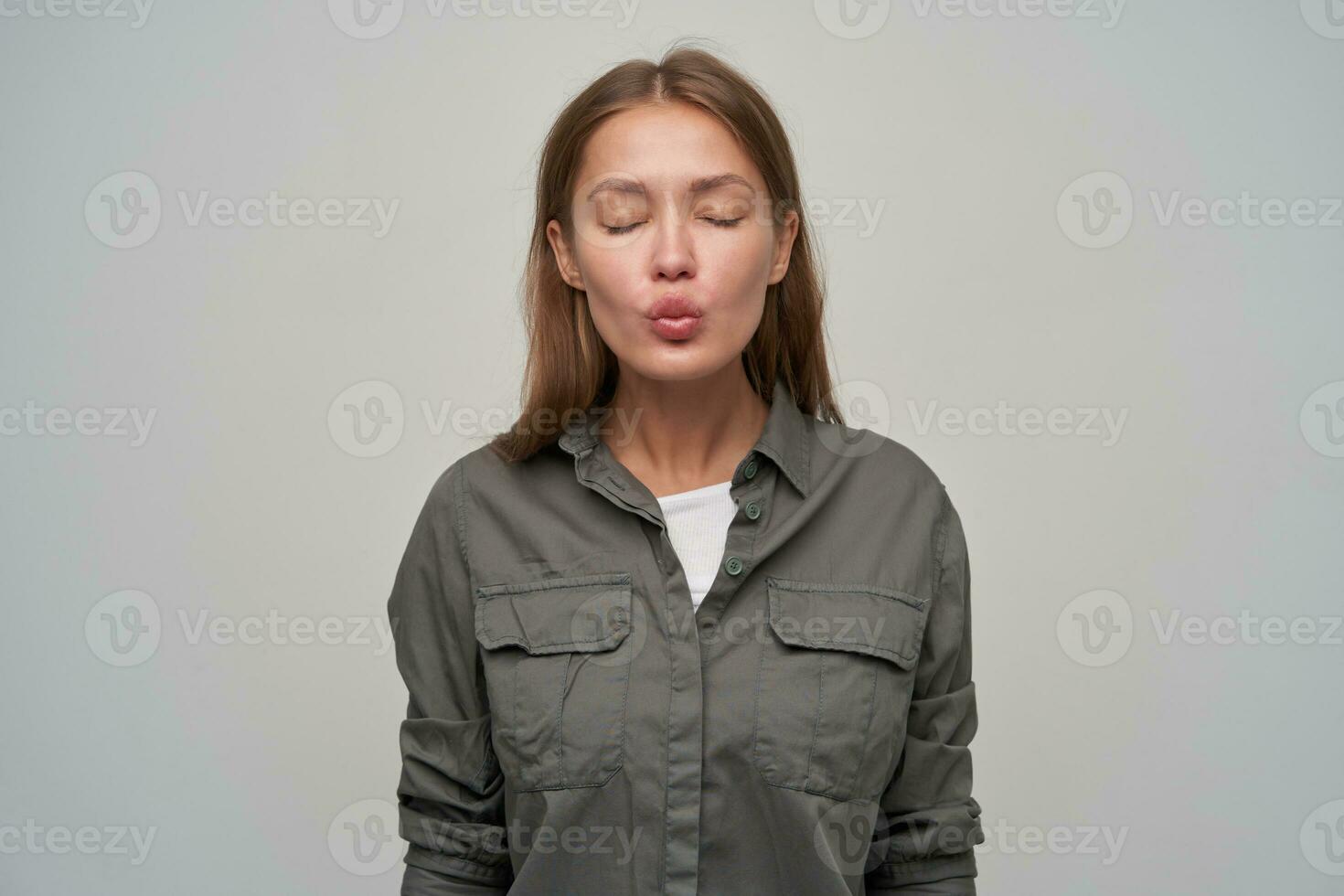 Portrait of attractive, adult european girl with brown long hair. Wearing grey shirt and dreaming. Lovely kissing with closed eyes. Natural. Stand isolated over grey background photo