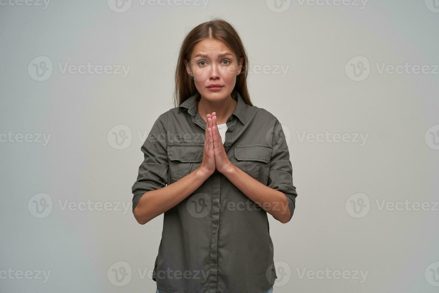 Teenage girl, unhappy woman with brown long hair. Wearing grey shirt and keep her palms together, asks for something. Watching at the camera isolated over grey background photo