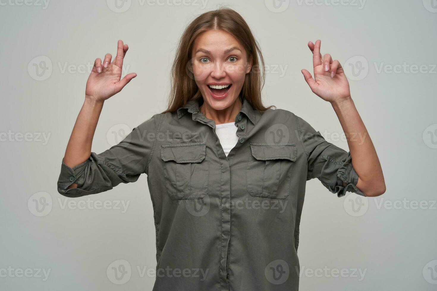 Young lady, pretty woman with brown long hair. Wearing grey shirt and crossed her fingers. Cheerful, making a wish. Smiling. Watching at the camera isolated over grey background photo