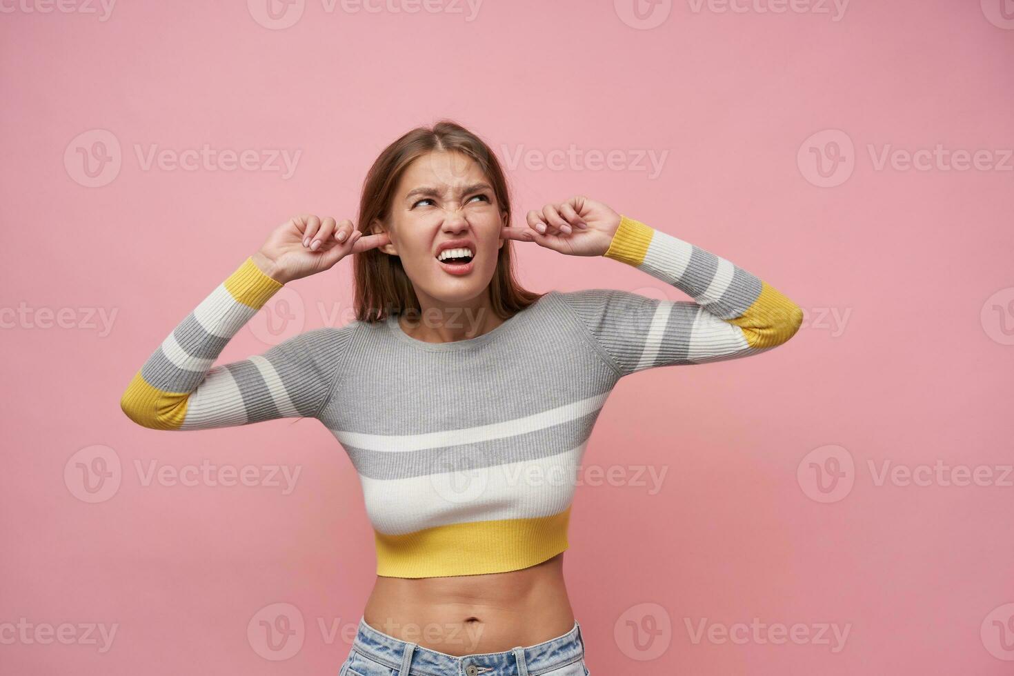 Portrait of attractive, adult girl with brown long hair. Wearing striped blouse and jeans. Close her ears and squints from the noise. Watching up at copy space, isolated over pastel pink background photo
