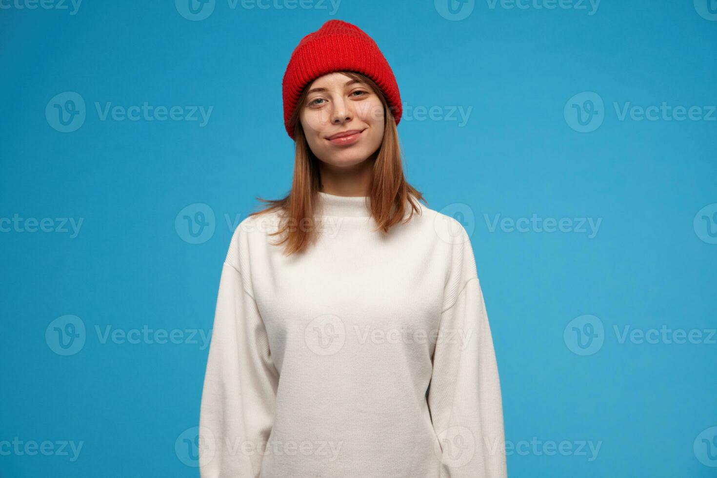 Relaxed looking woman, beautiful girl with brunette hair. Wearing white sweater and red hat. People and emotion concept. Watching at the camera and smile, isolated over blue background photo