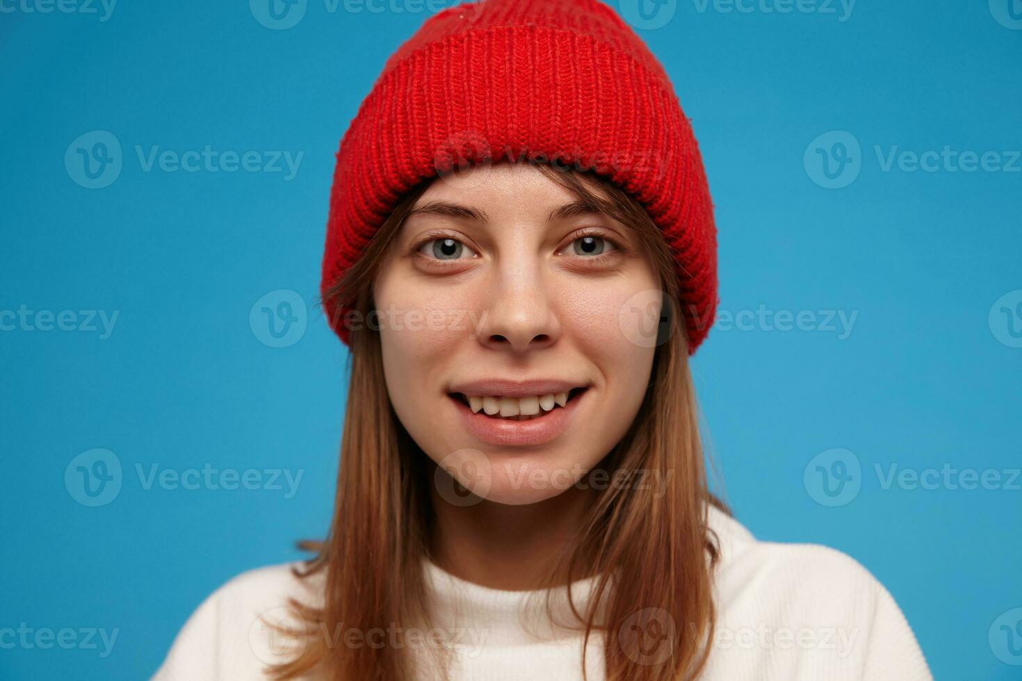 Cool looking woman, happy girl with brunette hair. Wearing white sweater and red hat. People and emotional concept. Watching at the camera with a smile, closeup, isolated over blue background photo