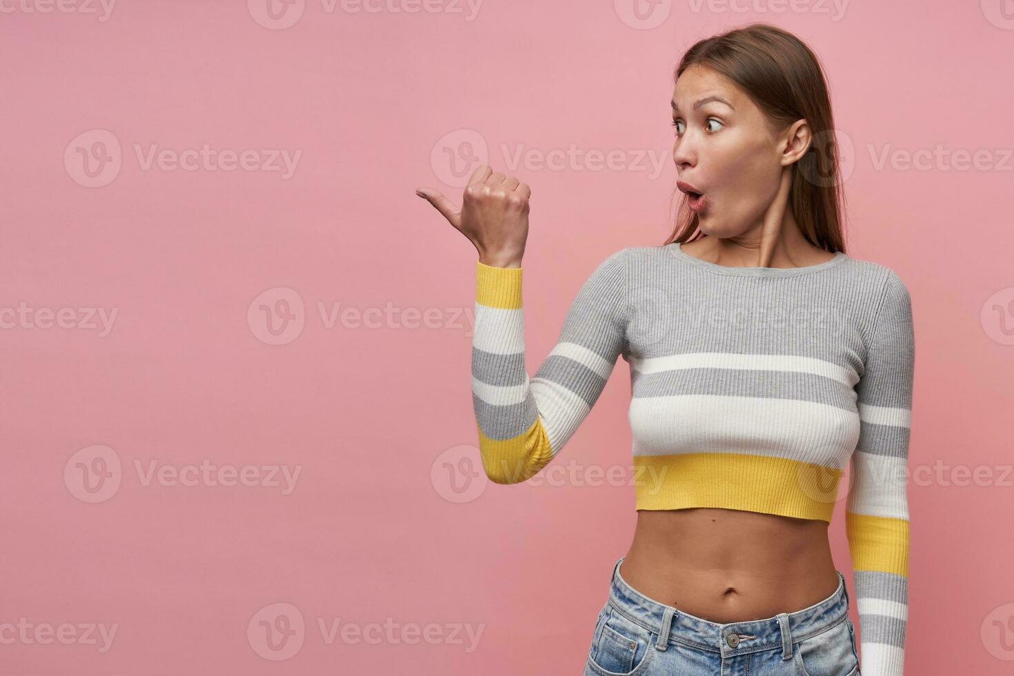 Young lady, pretty woman with brown long hair. Wearing striped blouse and jeans. Shocked, pointing with thumb and watching to the left at copy space, isolated, over pastel pink background photo