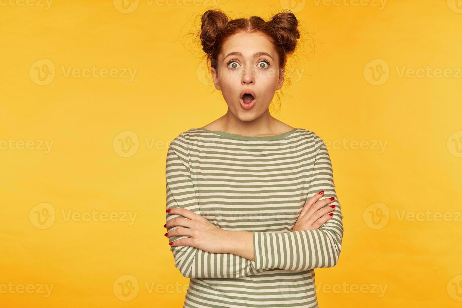 Teenage girl, shocked looking red hair woman with two buns. Wearing striped sweater and watching at the camera with hands crossed on a chest and surprised face. Stand isolated over yellow background photo