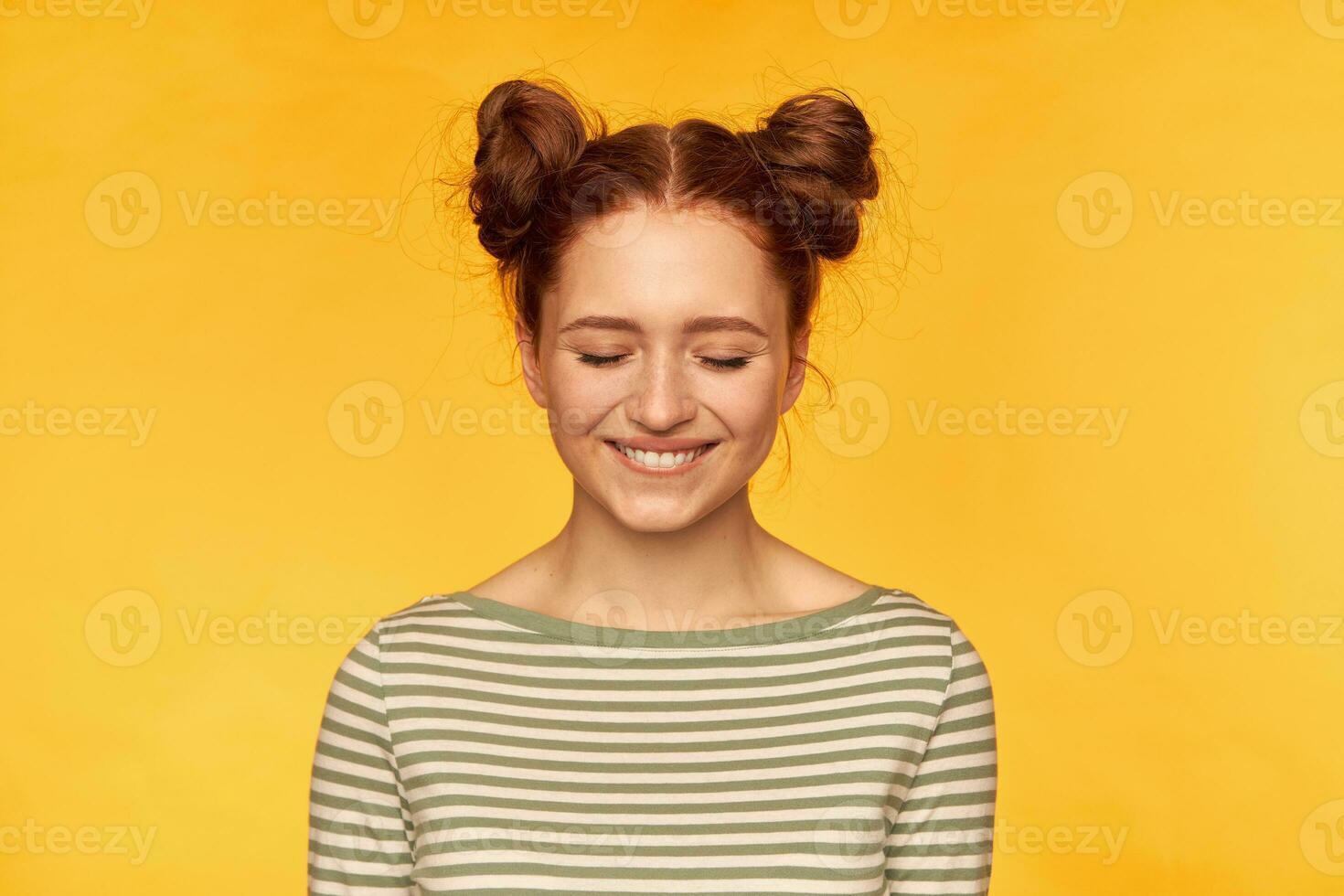 joven dama, bonito jengibre mujer con dos bollos vistiendo a rayas suéter y mirando entusiasmado. sensación muy contento a el momento. estar aislado terminado amarillo antecedentes con cerrado ojos foto