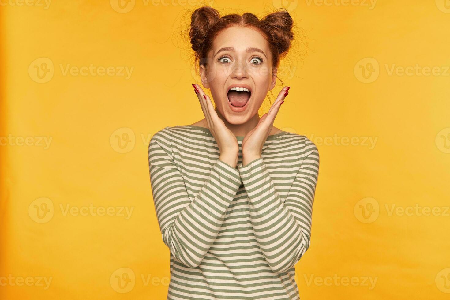 Young lady, horrified looking ginger woman with two buns. Shows how much she frightened of what she see. Wearing striped sweater and screams at the camera isolated over yellow background photo