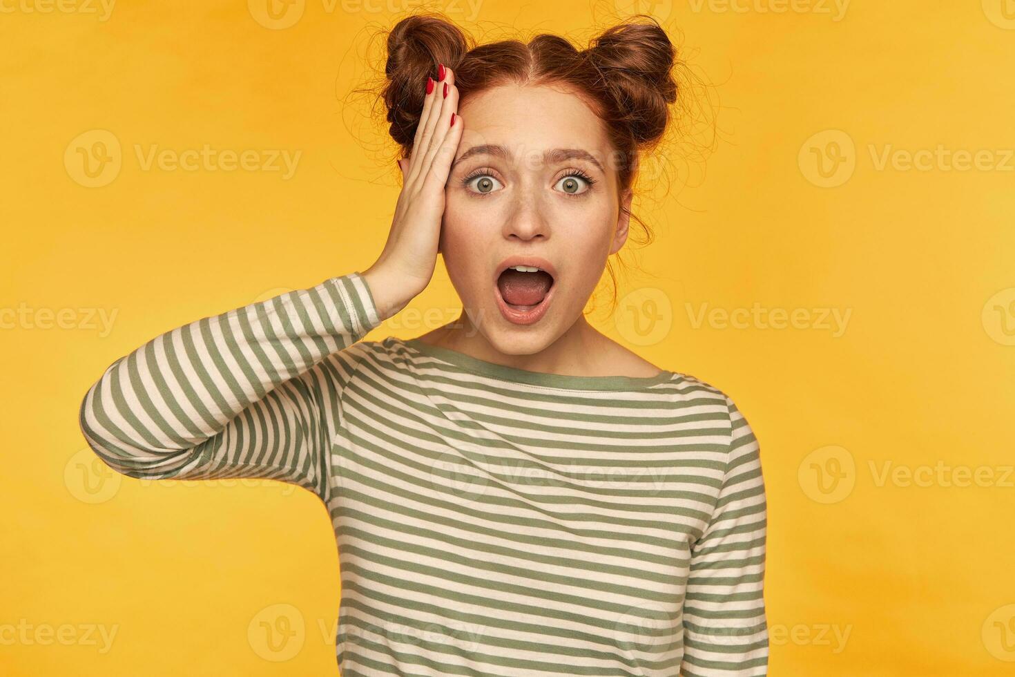 Portrait of attractive, excited red hair girl with two buns. Wearing striped sweater and hear shocking news, touching her head. Watching at the camera isolated over yellow background photo