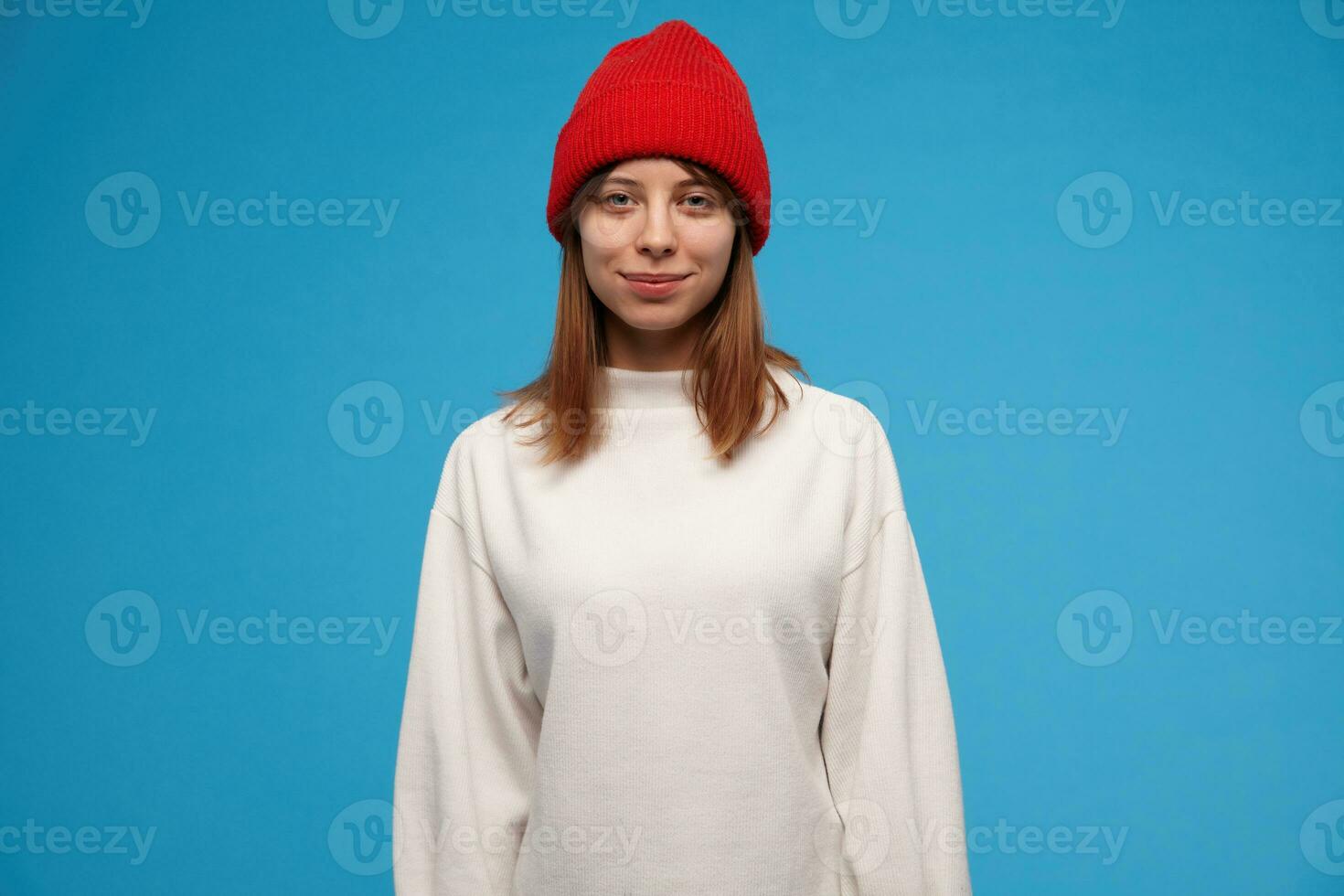 Teenage girl, cool looking woman with brunette hair. Wearing white sweater and red hat. People and emotional concept. Watching at the camera and smile, isolated over blue background photo