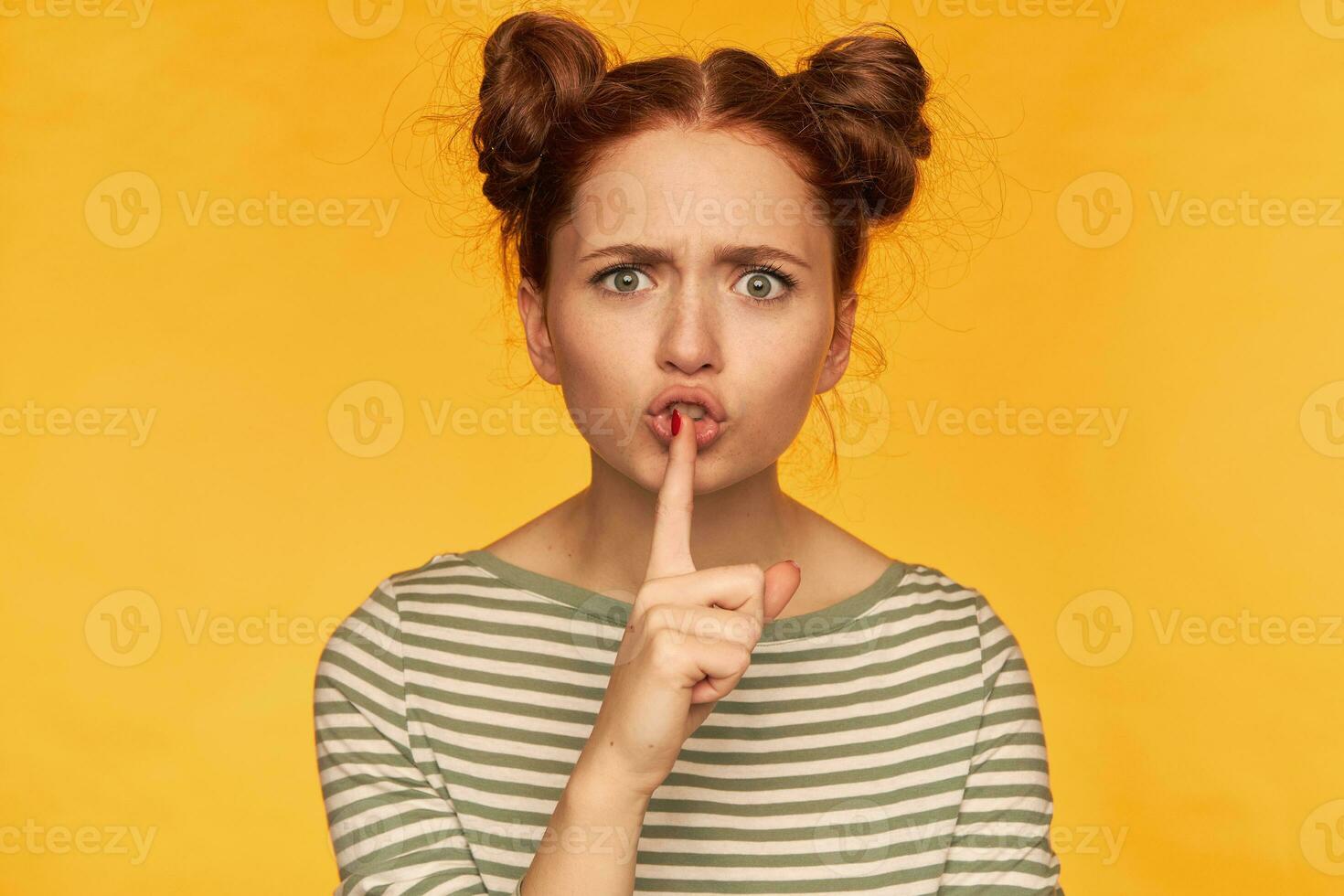 Portrait of attractive, adult girl with two buns and big eyes. Wearing striped blouse and showing silence sign, looking angry, unsatisfied. Watching at the camera isolated over yellow background photo