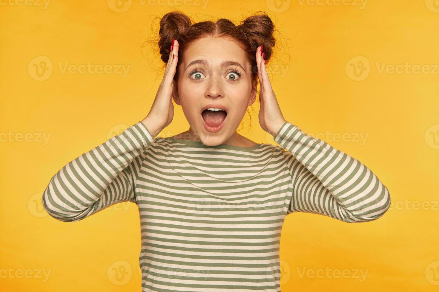 Young lady, emotional ginger woman with two buns. Wearing striped sweater and hear shocking news, touching her head with both hands. Watching at the camera isolated over yellow background photo