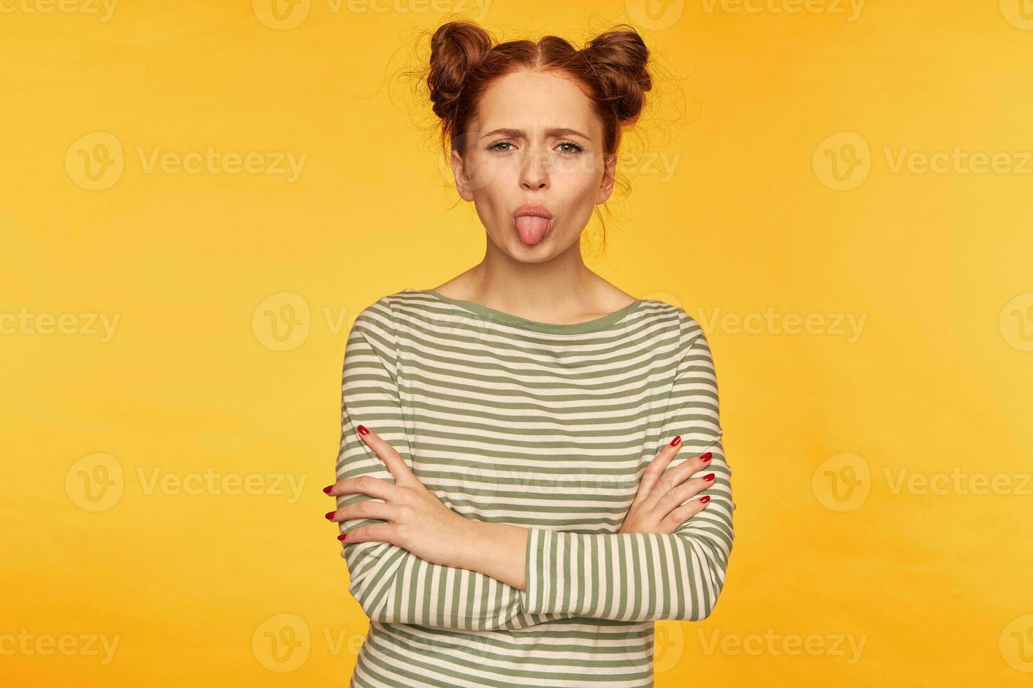 Portrait of attractive, red hair girl with two buns. Wearing striped sweater and showing a tongue to the camera with hands crossed on a chest. Stand isolated over yellow background photo