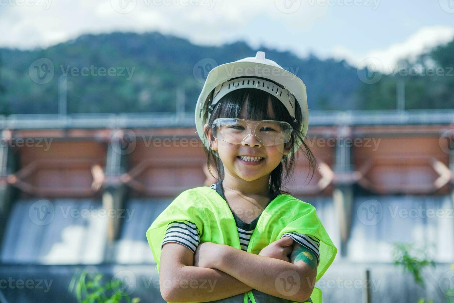 retrato de un pequeño niña ingeniero vistiendo un verde chaleco y blanco casco sonriente felizmente en el antecedentes de el represa. conceptos de ambiental ingeniería, renovable energía y amor de naturaleza. foto